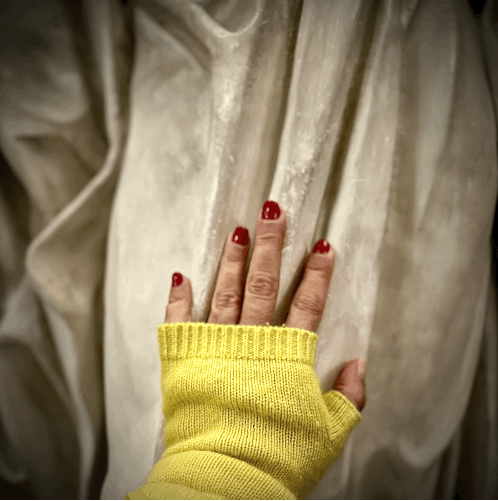 Main féminine posée sur une pierre claire, vêtue d’un gant jaune, symbole de contact entre le corps et la matière, entre le vivant et le sacré — Je marche donc je suis